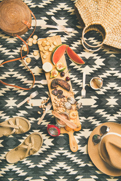 Summer picnic setting. Flat-lay of fresh fruit, smoked sausage, nuts, brie cheese, pate, cracker and woman straw accessories over linen blanket background, top view. Outdoor gathering or lunch