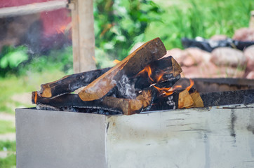 Brightly burning in metal box logs firewood for barbecue outdoor on sunny day. Orange flame and white smoke  and green grass background. Camping, safety and tourism concept.