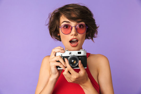 Pretty woman 20s in sunglasses smiling and taking photo on retro camera, isolated over violet background