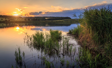 Sunset on the lake. The water reflects the setting sun and clouds. - obrazy, fototapety, plakaty