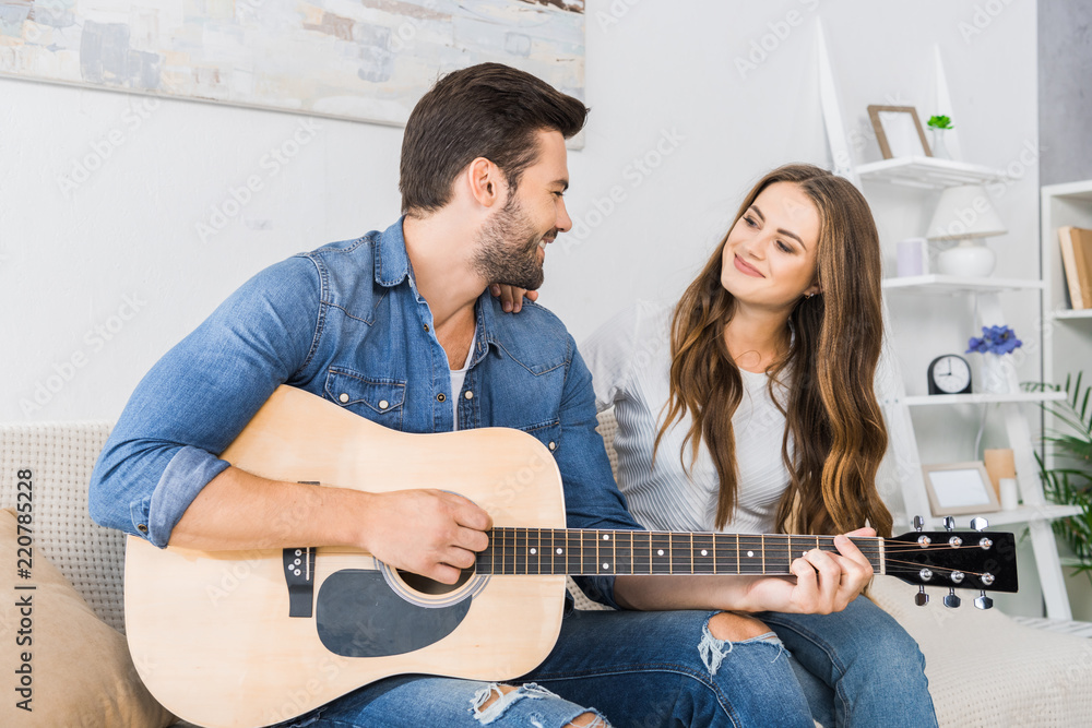 Wall mural happy young man playing on acoustic guitar and looking at his girlfriend on sofa at home