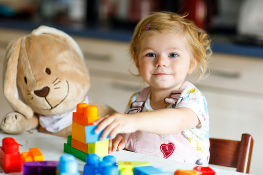 Adorable toddler girl with favorite plush bunny playing with educational toys in nursery. Happy healthy child having fun with colorful different plastic blocks at home. Cute baby learning creating.
