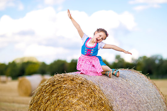 Cute little kid girl in traditional Bavarian costume in wheat field. German child with hay bale during Oktoberfest in Munich. Preschool girl play at hay bales during summer harvest time in Germany.