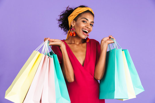 Image of african american woman 20s in hair band and earrings smiling and holding colorful paper shopping bags with purchases, isolated over violet background