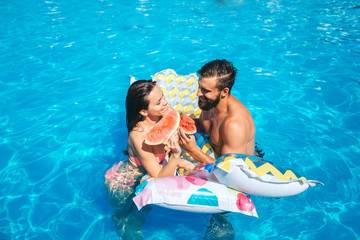 A picture of couple swimming in pool and leaning to air mattress. They look at each other. Also couple has pieces of watermelon in hands. They eat it.
