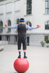 The clown juggles with small balls, standing on a large ball in the street of a European city - 220788620