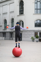 The clown juggles with small balls, standing on a large ball in the street of a European city