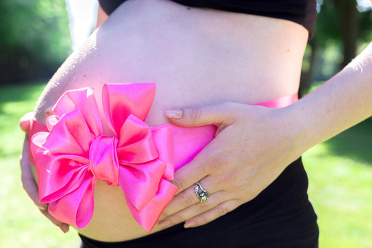 A young pregnant girl holds herself by the belly, which is tied with a festive pink ribbon with a bow