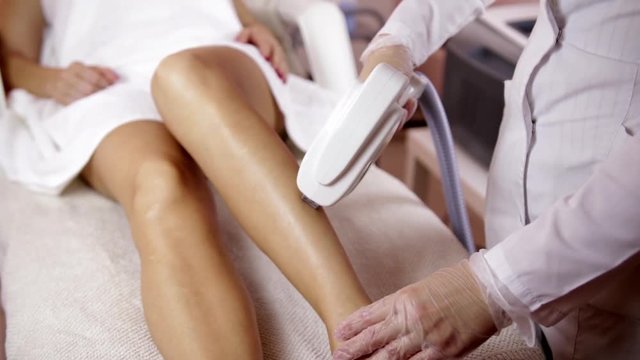 Laser hair removal on ladies legs. The girl is lying on the couch in the medical glasses in the treatment room. The hands of the cosmetologist make the epilation of the legs