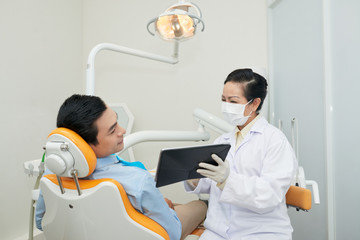 Woman in mask and gloves showing tablet to Asian man in dental chair while working in clinic