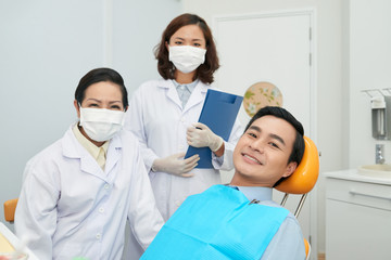 Asian man in dental chair smiling at camera with coworking doctors in white medical gowns standing near