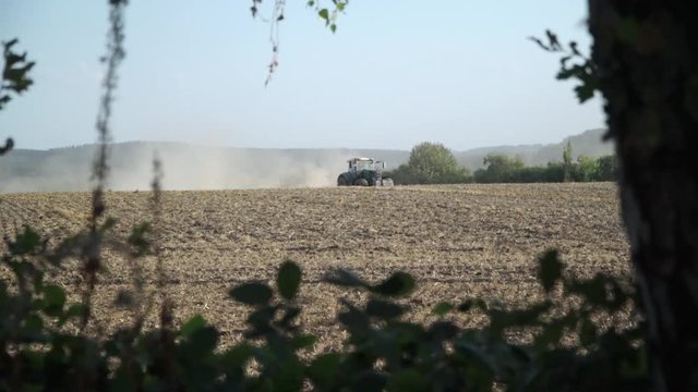 Traktor f&auml;hrt &uuml;bers Feld und wirbelt Staub auf, Baum und Geb&uuml;sch im Vordergrund