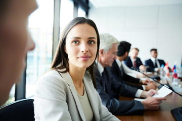 Head and shoulders portrait of young businesswoman looking at colleague while sitting at meeting table in conference room, copy space