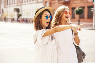 Fashion portrait of two young stylish hippie brunette and blond women models in summer sunny day in white hipster clothes posing on the street background. Pointing on shop sales