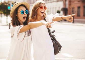 Two fashion young stylish hippie brunette and blond women models in summer sunny day in white hipster clothes posing on the street background. Pointing on shop sales