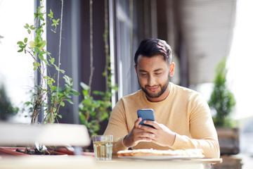 Contemporary guy with smartphone texting or reading message while sitting by served table by lunch in cafe