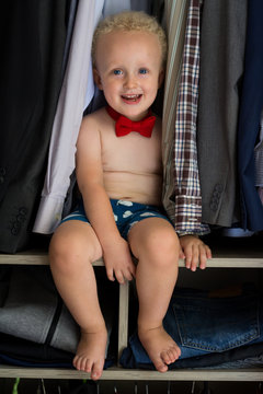 Little gentleman is getting ready. Cute and handsome boy with curly blond hair wearing underpants and red bow tie, sitting and smiling in dad's wardrobe.