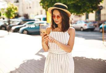 Portrait of happy teen girl in summer hipster clothes listening music on her headphones from smartphone in the street on a sunny day