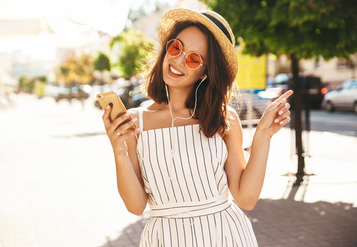 Portrait of happy smiling teen girl in summer hipster clothes listening music on her headphones from smartphone in the street on a sunny day