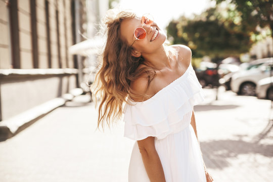 Beautiful cute smiling blond teenager model with no makeup in summer hipster white dress running on the street in sunglasses. Turn around