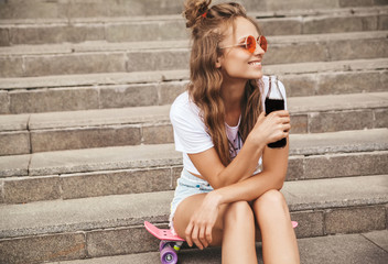 Beautiful cute blond teenager model in summer hipster white clothes sitting on pink penny skateboard on stairs. Drinking soda from bottle - obrazy, fototapety, plakaty