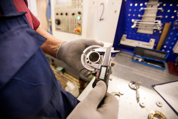Gloved worker of factory measuring diameter of metallic detail with calipers by workplace