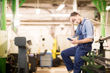 Embarrassed young engineer of factory reading notification in smartphone and touching his forehead