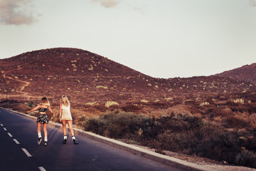 couple of beautiful blonde girls viewed from rear skating and moving on an asphalt road during a...