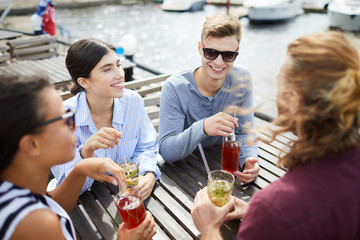 Cheerful young friends having rest in outdoor cafe and having cocktails by talk on sunny day