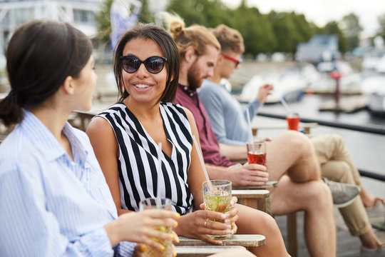 Young woman in sunglasses talking to one of her friends while having drinks and relaxing outdoors
