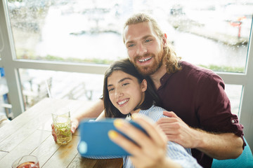 Young cheerful couple making selfie while sitting by table in cafe and having drinks