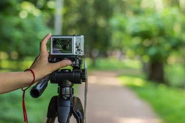 photography hobby and equipment of human hand and fingers on small gray cheap digital camera stay on tripod on unfocused blurred bokeh natural outdoor park environment, copy space