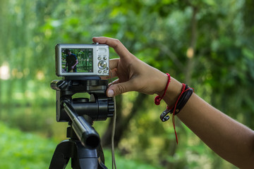 photography hobby and equipment of human hand and fingers on small gray cheap digital camera stay on tripod on unfocused blurred bokeh natural outdoor park environment, copy space