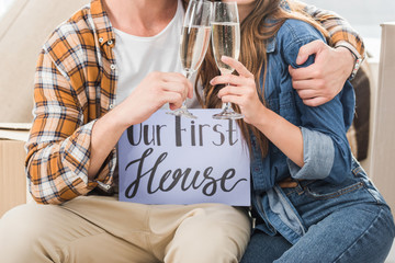 partial view of couple with our first house card clinking glasses of champagne, moving home concept
