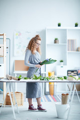 Young woman in workwear making bunch of green leaves and flowers while standing by desk