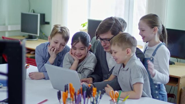 Group of interested multiethnic schoolchildren smiling and looking at laptop computer screen while watching something with young male teacher