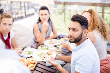 Portrait of frowning Middle-Eastern man complaining about food during lunch with friends in cafe,...