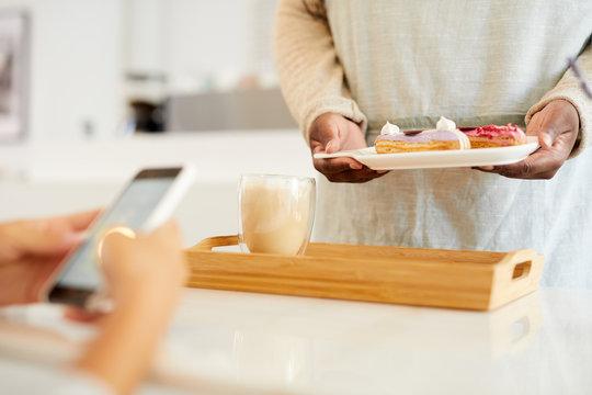 Wooden tray with milkshake on table and hands of waitress holding plate with cakes over it