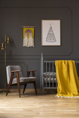 Grey armchair next to bed with yellow blanket in baby's bedroom interior with poster. Real photo