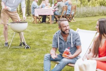 Tuinposter Shashliks being put on a grill by a man during a BBQ party. Blurred foreground with a woman and a man sitting on deckchairs and talking. Other people by a table in the background. © Photographee.eu