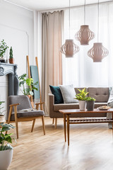 Lamps above wooden table with flowers in flat interior with grey armchair and sofa. Real photo