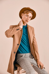 Young male model with red hair and freckles posing in brown coat and hat, plaid pants, turquoise...