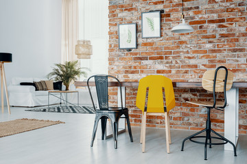 Modern chairs at dining table against red brick wall with posters in bright apartment interior. Real photo