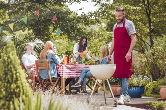 A young man wearing a burgundy apron cooking on a white grill. People sitting around a table and having fun during a celebration in the backyard.