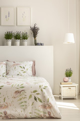Lamp above cabinet next to bed with plants on headboard in feminine bedroom interior. Real photo