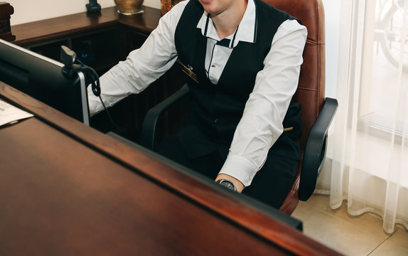 The Administrator Is Sitting At The Hotel Reception Desk. Business Photo Of The Man Employee. Young Guy Receptionist In The Black Uniform Is Working At The Computer Screen Laptop.