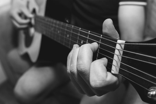 Man at home is playing the guitar. Guys hands are taking the chord on strings. Music making lifestyle concept. Free time hobby for everyone. Retro black and white guitar photo.
