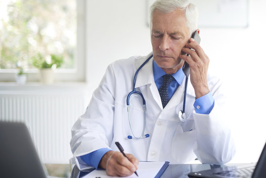 Portrait of male doctor making call with his patient and writing down the diagnosis while sitting at consulting room.