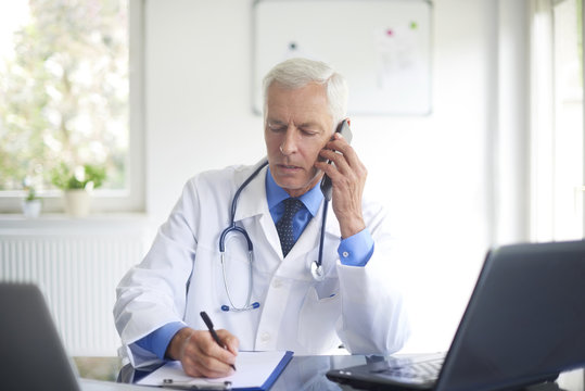Portrait of male doctor making call with his patient and writing down the diagnosis while sitting at consulting room.