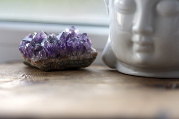 Purple and gemmy amethyst stone with White Buddha head on the windowsill background. Rough ametist crystals on home altar, spiritual ritual meditation place. Religion symbol